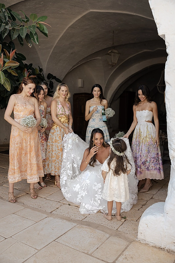 Bride with bridesmaids during a bridesmaids reveal moment, holding baby's breath bouquets in a stone courtyard with arched walkway and lanterns