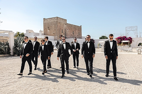 Groomsmen portrait of men in black tuxedos with sunglasses walking in a gravel courtyard by white walls and agave plants under blue sky