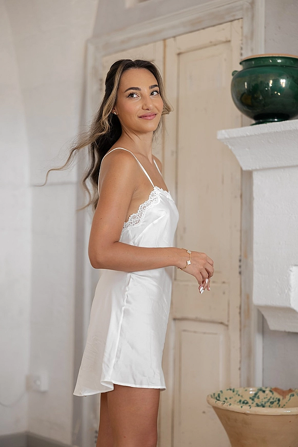 Bridal getting ready portrait of the bride getting ready outfit in a white satin slip dress with lace trim, by a white mantel in a bright room