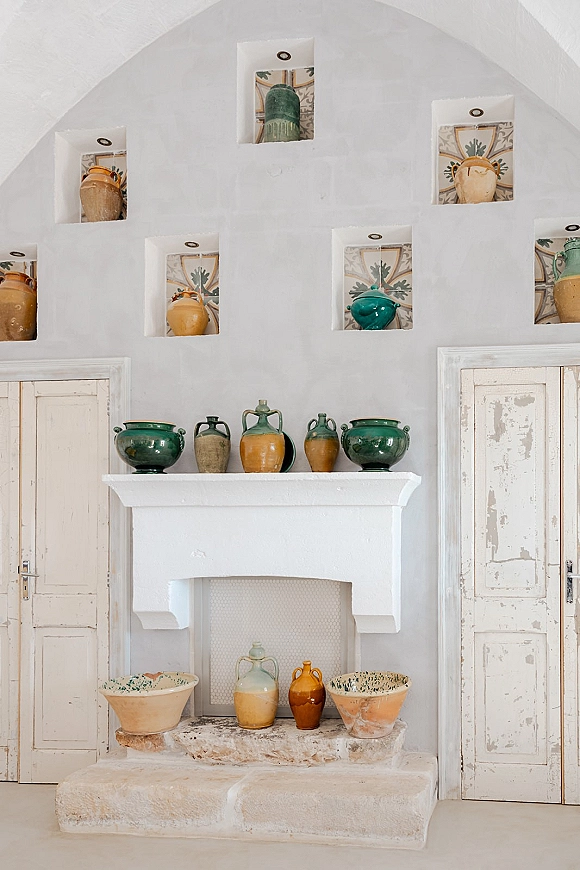 Venue interior with white plaster arch wall, featuring ceramic pottery display in wall niches above a stone hearth and distressed doors
