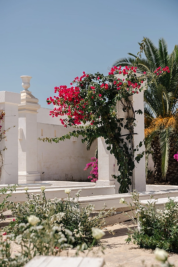 Outdoor ceremony decor with bougainvillea wedding arch framing white stucco pillars and benches, set against palm trees and blue sky