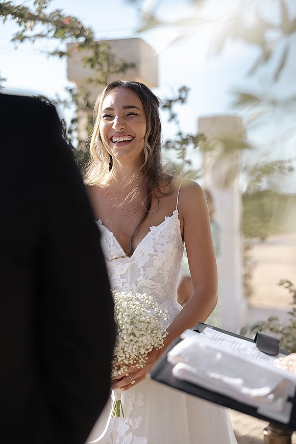 Ceremony moment as the bride laughs, holding a baby’s breath bridal bouquet in a lace spaghetti-strap dress outdoors by stone and greenery