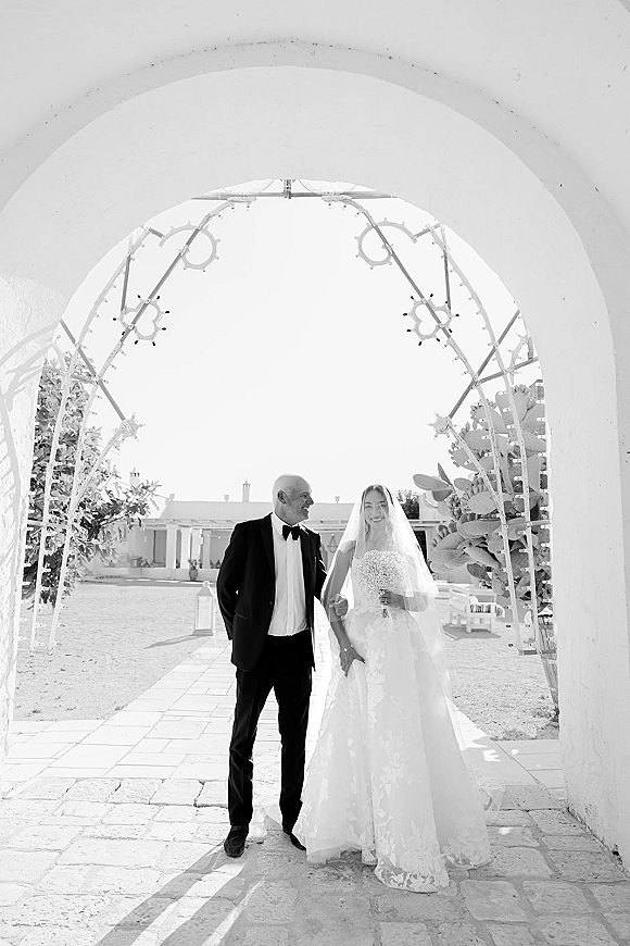 Couple portrait of bride in lace wedding dress and veil holding a white bouquet beside groom in tuxedo under a white archway courtyard walkway