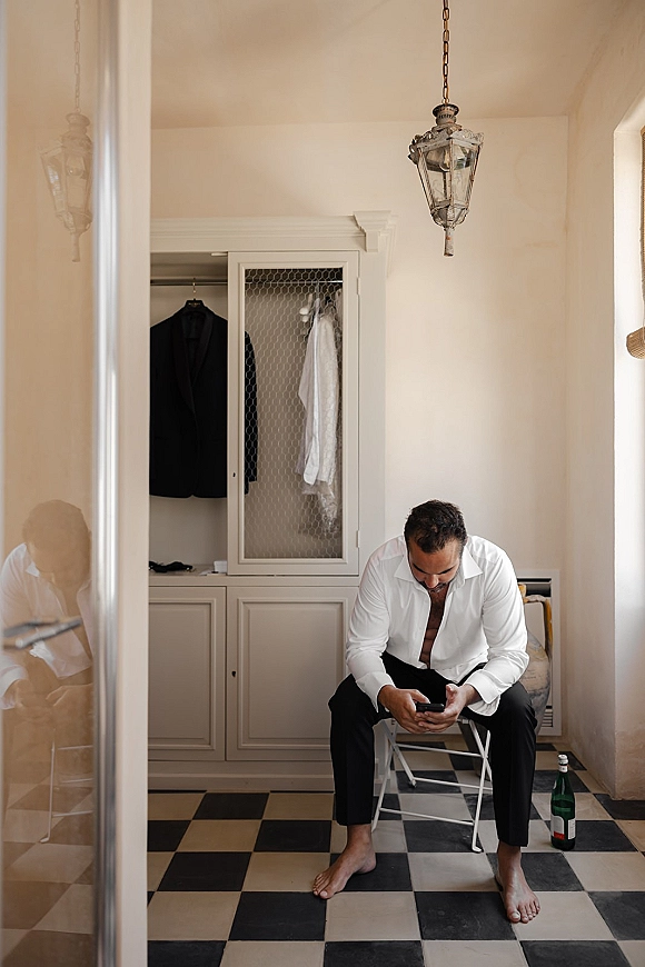 Groom getting ready, groom on phone while sitting in a white dress shirt and black trousers in a dressing room with checkered tile floor