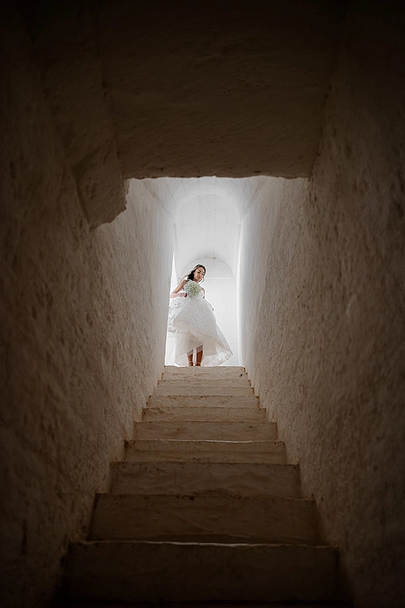 Bridal portrait of a bride on stairs holding a minimal bouquet, looking down in a strapless dress by an arched doorway and white walls