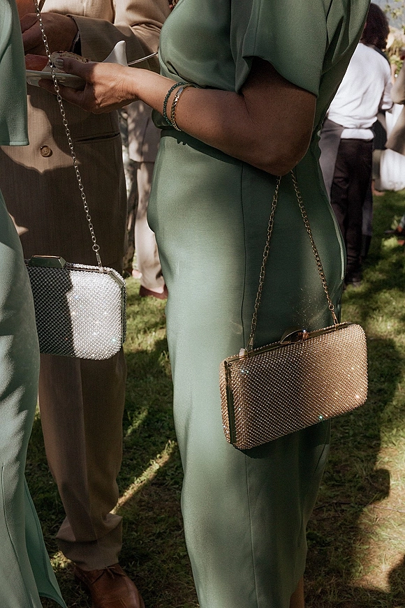 Wedding guest outfit in a sage green dress holding a rhinestone clutch and plate of food on a sunny lawn with trees and guests behind