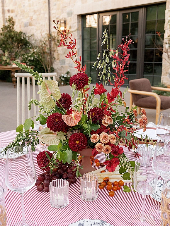 Wedding centerpiece with a tall wedding centerpiece of red dahlias and anthurium in a pedestal vase on a striped table with candles and grapes in a stone courtyard