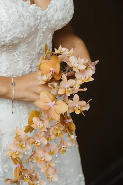 Bridal bouquet with a cascading orchid bouquet in peach tones held against an off-the-shoulder beaded lace gown in a dark indoor setting