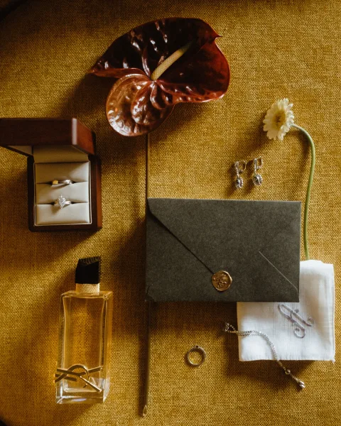 Wedding flat lay with wedding invitation flat lay, ring box, earrings, perfume bottle, wax seal envelope on gold textured fabric with anthurium
