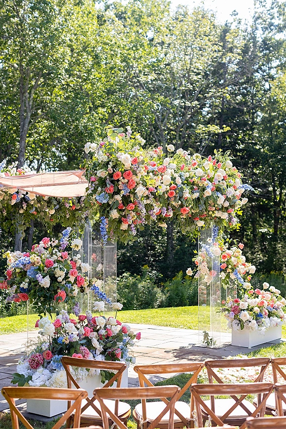 Wedding ceremony arch with pastel flowers and roses on clear acrylic columns, lined by aisle blooms on a sunlit lawn patio