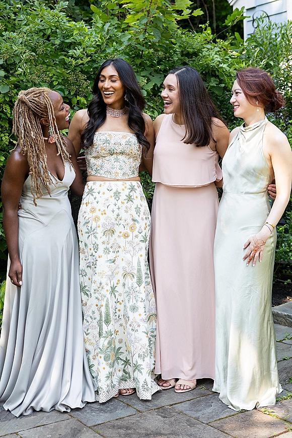 Bridesmaid group photo of friends laughing together in mismatched satin dresses, standing by a greenery hedge on a stone patio