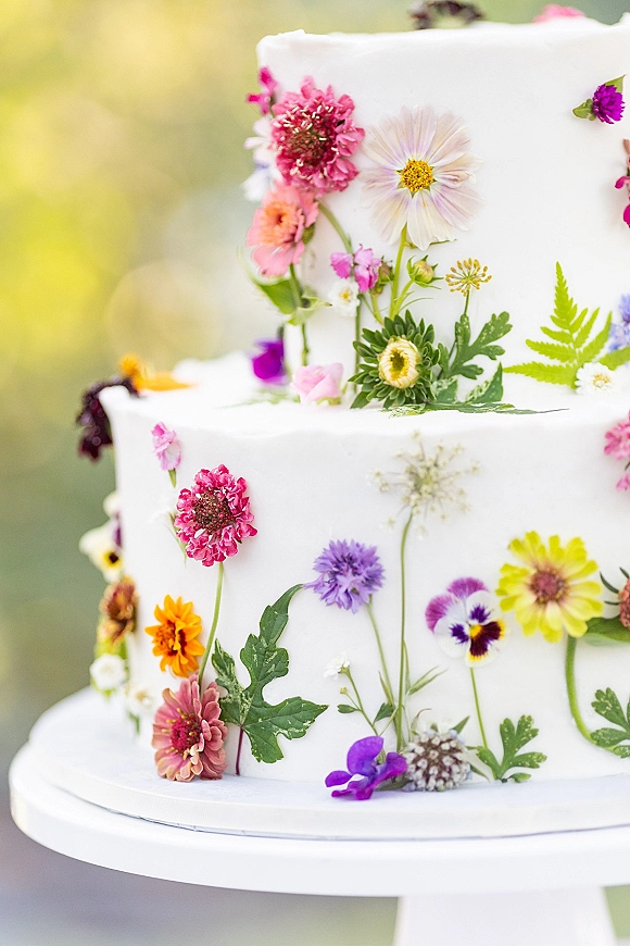 Wedding cake with wildflower wedding cake blooms and greenery on a stand, white frosted tiers set against blurred outdoor greenery