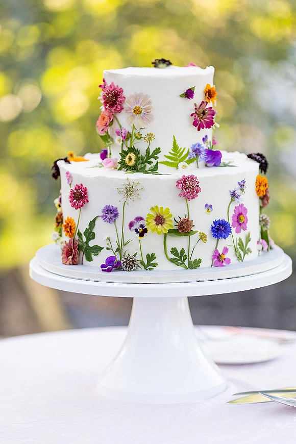 Wedding cake with pressed flower details, a two-tier white buttercream design on a stand against blurred outdoor greenery and trees