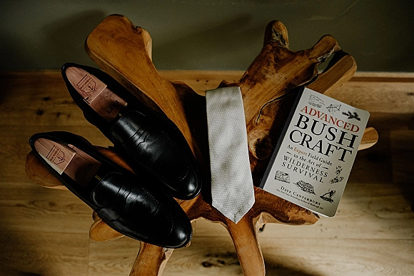 Groom accessories flatlay with black leather dress shoes, white tie, and a book on a wood accent table over a wood floor