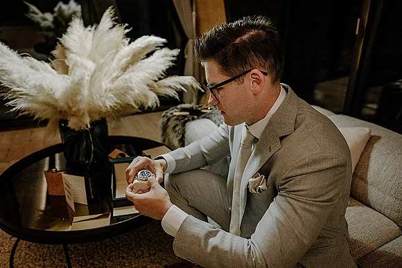 Groom getting ready, groom checking watch in a light gray suit with glasses, standing by a lounge sofa with pampas grass decor
