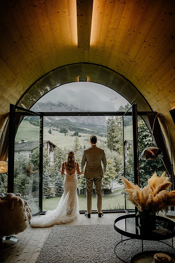 Couple portrait of bride and groom from behind holding hands, her lace dress train flowing by glass doors facing mountains