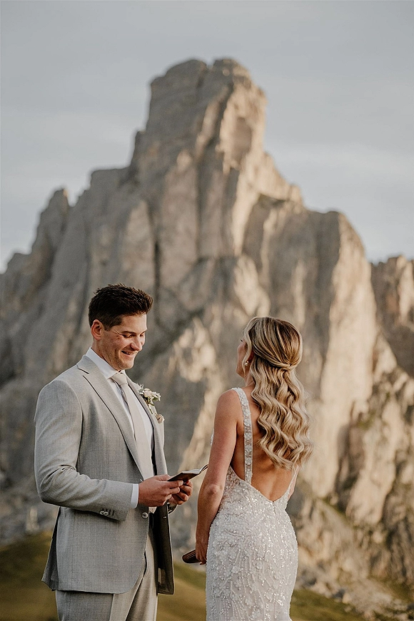 Wedding vows as the groom reads from a vow booklet beside the bride in a backless dress, with mountain peaks and rocky landscape behind