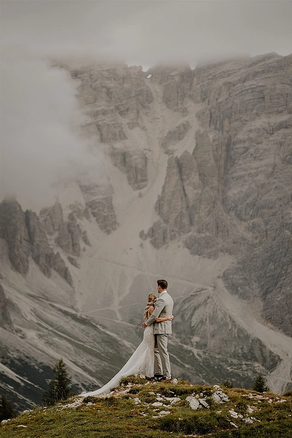 Couple portrait of bride in a long-train wedding dress and groom in suit embracing on a rocky ridge above foggy alpine cliffs