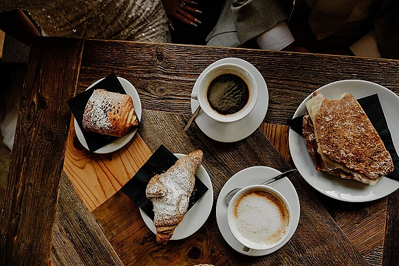 Wedding brunch spread with coffee cups, pastry and sandwich on a rustic wood table, black napkins and dessert plates in an indoor seating area