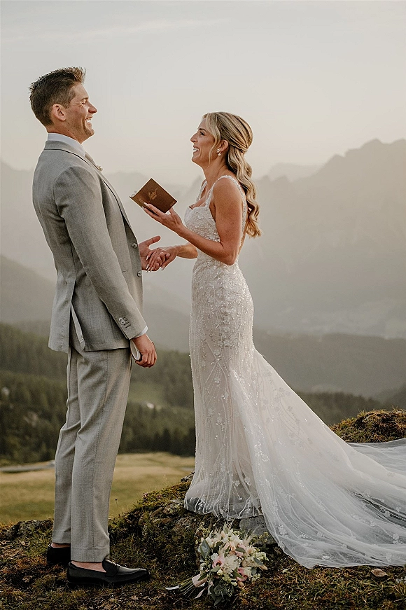 Wedding vows as bride reads from a vow book, holding hands with groom in a light gray suit on a mountain hillside under open sky