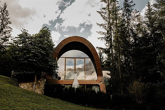 Wedding dress display in a cabin window, gown on hanger with a mountain reflection, forest trees and cloudy sky behind