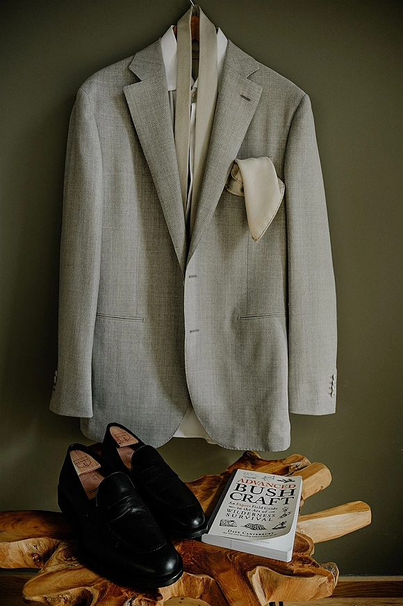 Groom suit details with a light gray wedding suit on a wooden hanger, beige tie and pocket square laid beside black loafers on a wood table, green wall behind
