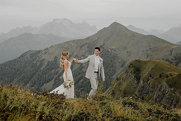 Couple portrait on a mountain ridge, bride in a white lace dress holding a bouquet beside groom in a suit under an overcast sky