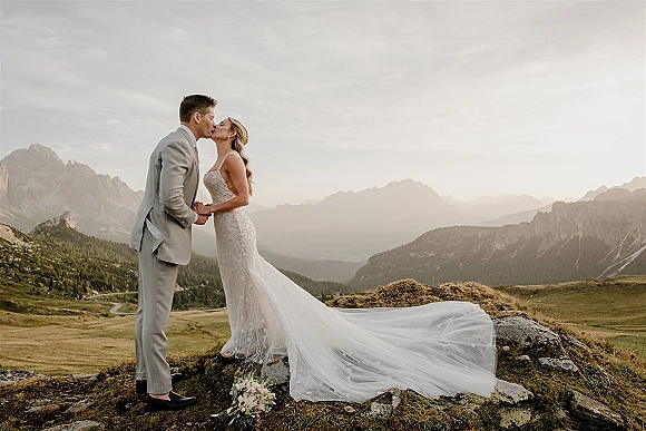 Wedding kiss as bride and groom embrace on a rocky mountain overlook, her long train and veil flowing, bouquet nearby against distant peaks
