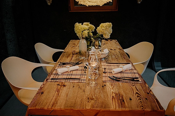 Reception tablescape with rustic wedding table setting on a wood table, woven placemats, glass vases of white flowers, wine glasses, and flatware.