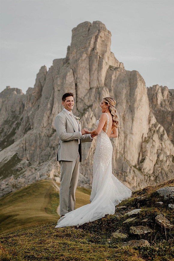 Couple portrait on a grassy mountainside, bride in a long-train wedding dress holding hands with groom in suit by rocky peaks
