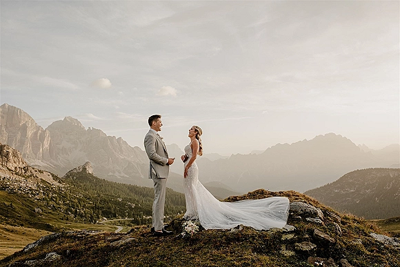 Couple portrait on a rocky mountain hilltop, bride in a long-train wedding dress facing groom in suit under cloudy sky