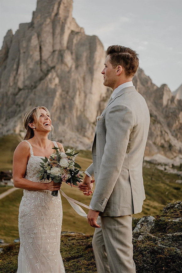 Couple portrait holding hands, bride with bouquet in a beaded wedding dress beside groom in a gray suit against mountain peaks and cliffs