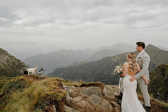 Couple portrait mountain wedding portrait from behind, bride in lace dress holding bouquet with ribbon on rocky cliff, goats below under clouds