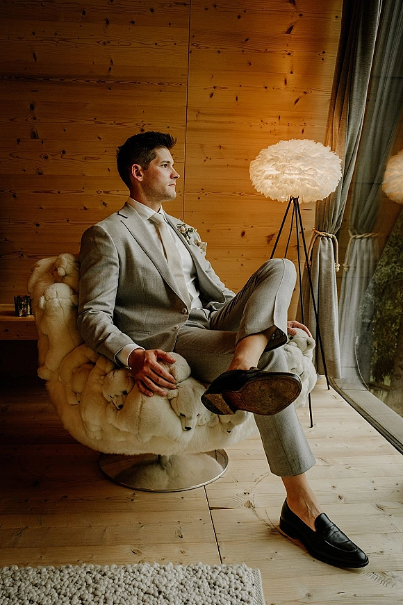 Groom portrait of a man seated in a furry armchair wearing a light gray suit with beige tie and boutonniere by a window in a wood-paneled room