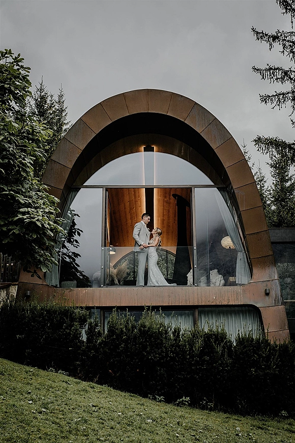 Couple portrait of a bride and groom on balcony, holding hands by a glass railing, framed by a modern arched building under overcast sky