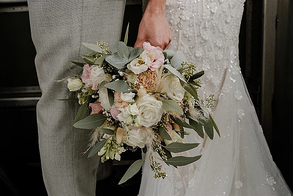Bridal bouquet of white roses and blush blooms with eucalyptus and pampas grass, held beside a dark wooden door with beaded dress detail