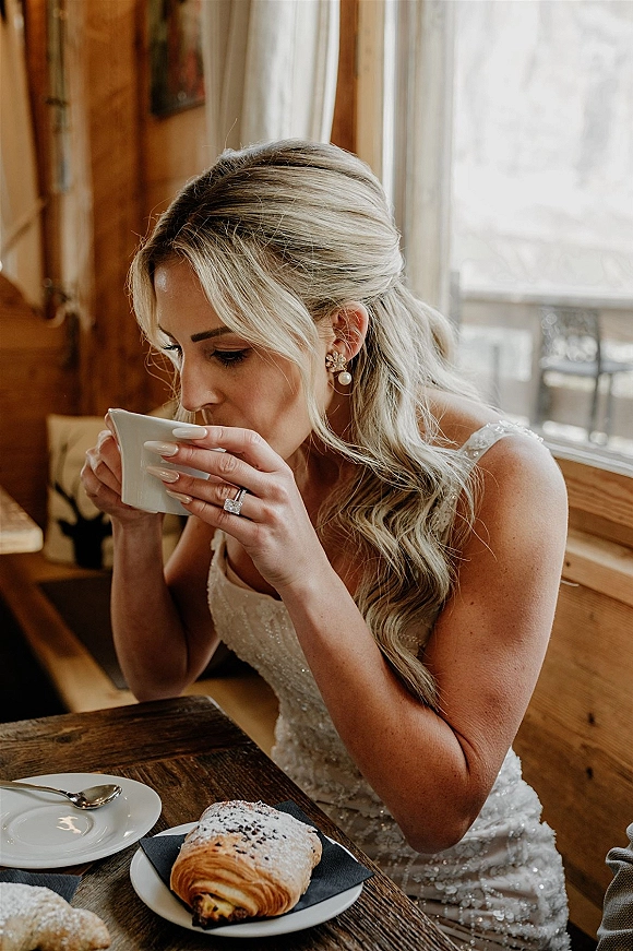 Bride portrait in a beaded wedding dress sipping coffee at a wooden table, showing engagement ring beside a pastry in rustic interior