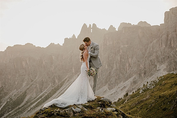 Wedding couple portrait of bride and groom kissing on a rocky cliff, veil and bouquet flowing with a dramatic mountain range behind