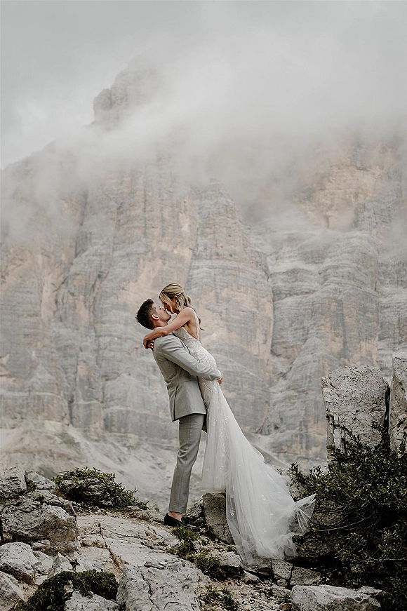 Wedding kiss portrait of groom lifting bride in a lace gown with long train on rocky cliffside amid foggy mountain cliffs