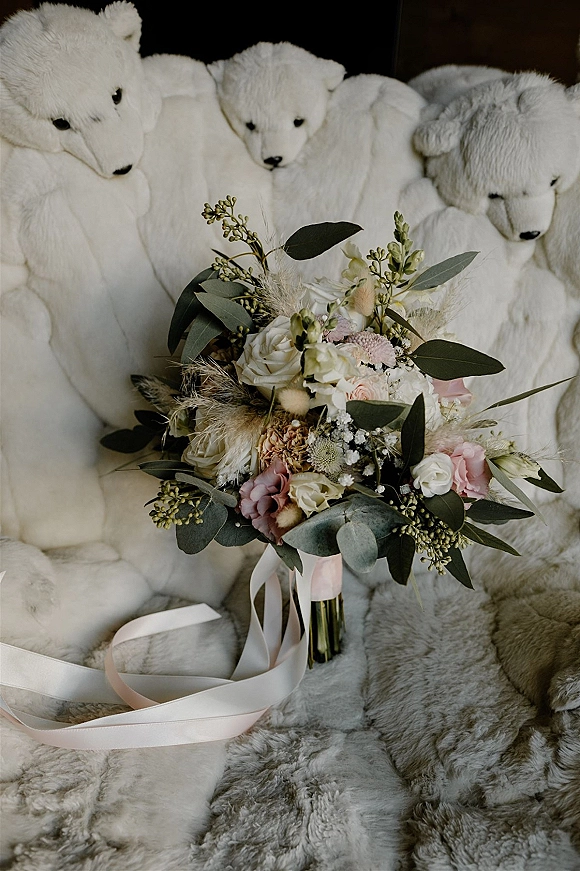 Bridal bouquet of white and blush roses with eucalyptus accents and baby’s breath, tied with ribbon on white faux fur beside teddy bears
