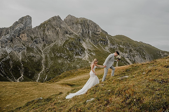 Couple portrait on a grassy alpine hillside, groom in light gray suit leading bride in a long-train wedding dress uphill toward rocky peaks