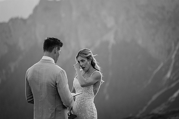 Wedding vows as bride wipes tears while groom reads from a vow book, wedding rings visible against a mountain landscape backdrop