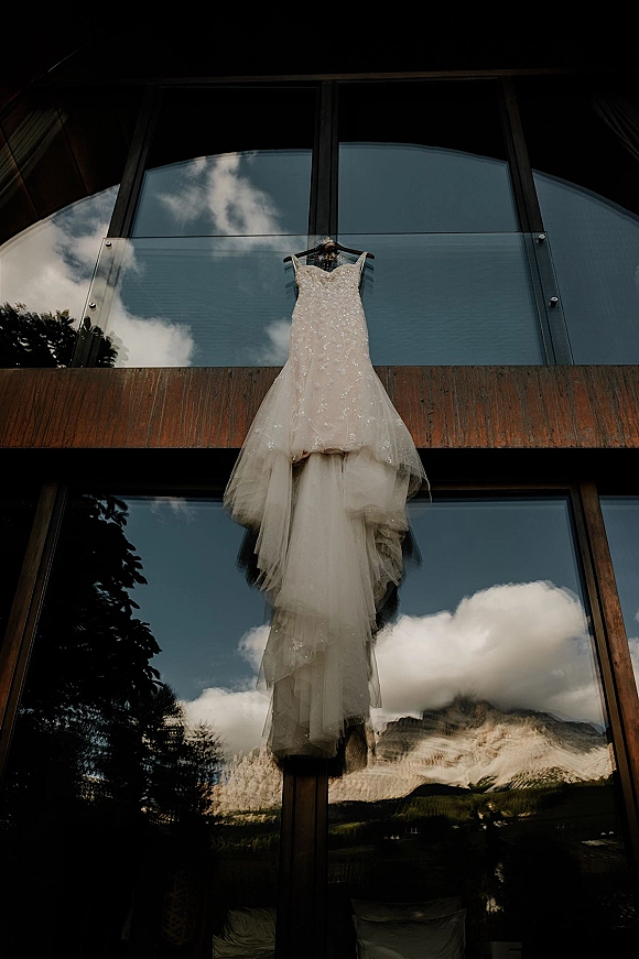 Wedding dress hanging in a window, beaded spaghetti straps over a tulle skirt, with mountain and cloud reflections in the glass