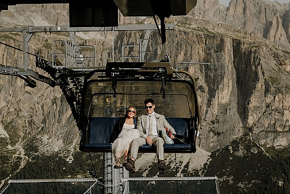 Couple portrait on a chairlift wedding photo, bride in a wedding dress and hiking boots beside groom in suit and sunglasses over mountain cliffs