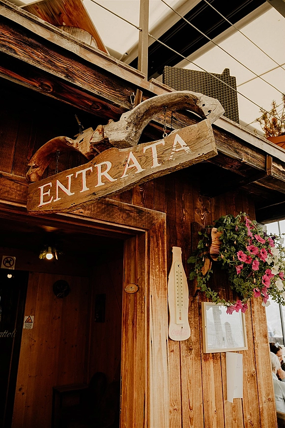 Wedding entrance sign hanging on chains, a rustic wedding signage detail with a pink flower basket by a wood cabin doorway under a canopy tent