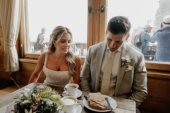 Reception moment as bride and groom at table share coffee and sandwiches at a rustic cabin wedding reception by a window with guests outside