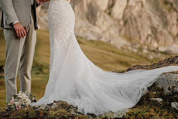 Wedding dress train flowing over rocks with beaded lace detail beside groom in gray suit and blush bouquet on a mountain hillside