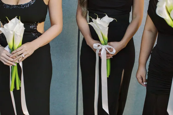 Bridesmaid bouquets of white calla lilies with long ribbon, held by women in black dresses with sequin detail against a gray wall