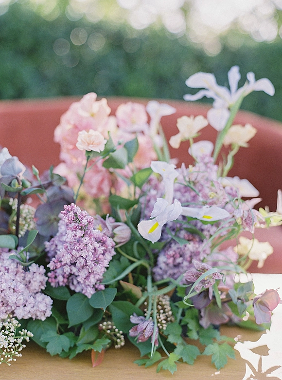 Wedding centerpiece with lilac flowers and irises in a vase, accented by blush blooms and greenery against soft garden bokeh lights