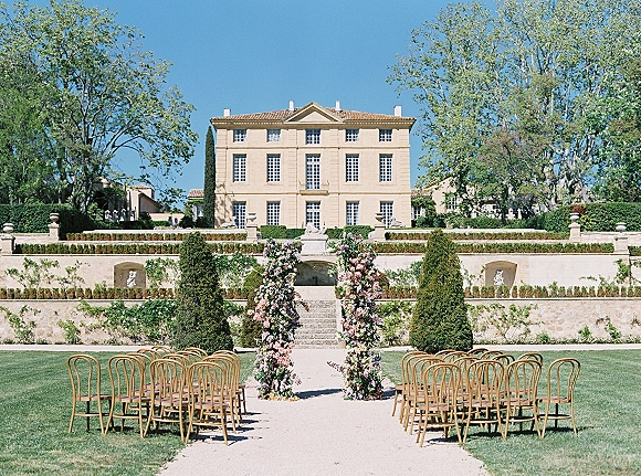 Ceremony setup for an outdoor wedding ceremony with wooden chairs and an aisle runner leading to a floral arch on an estate lawn under blue sky